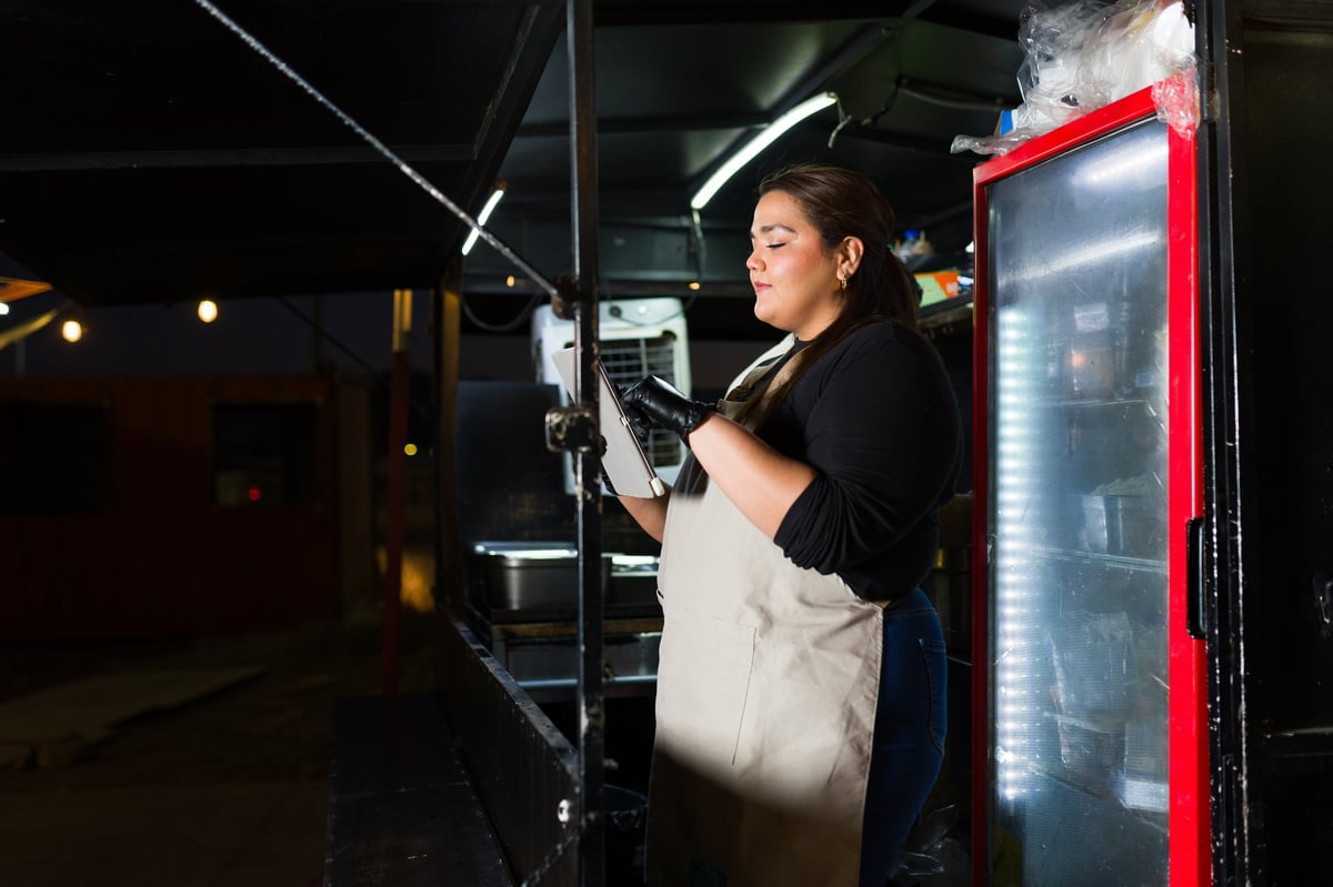 Hispanic woman wearing black gloves and apron using a tablet in a food truck at night, managing orders and preparing fast food