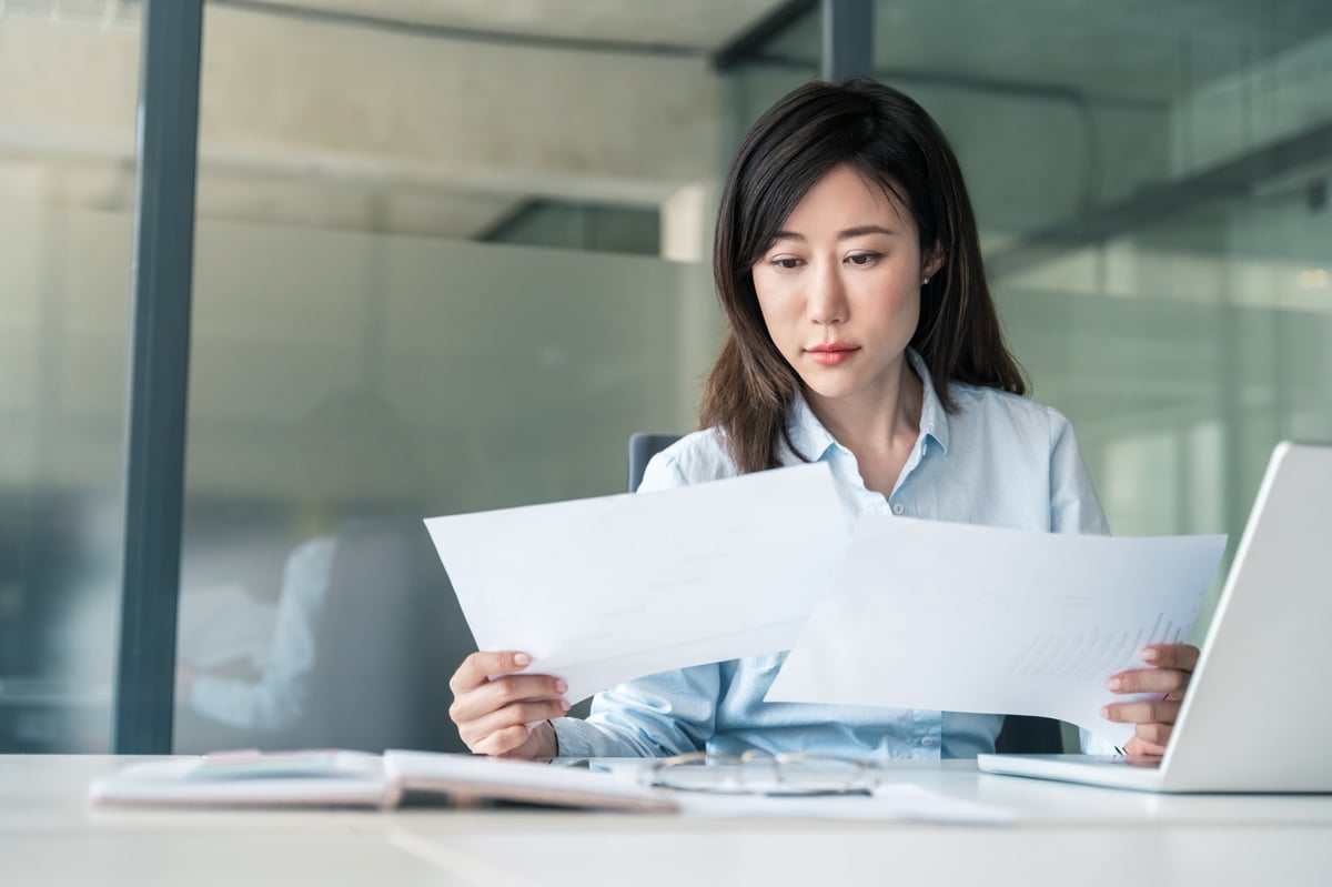Thinking Asian young business woman working on laptop computer reading financial document in office.