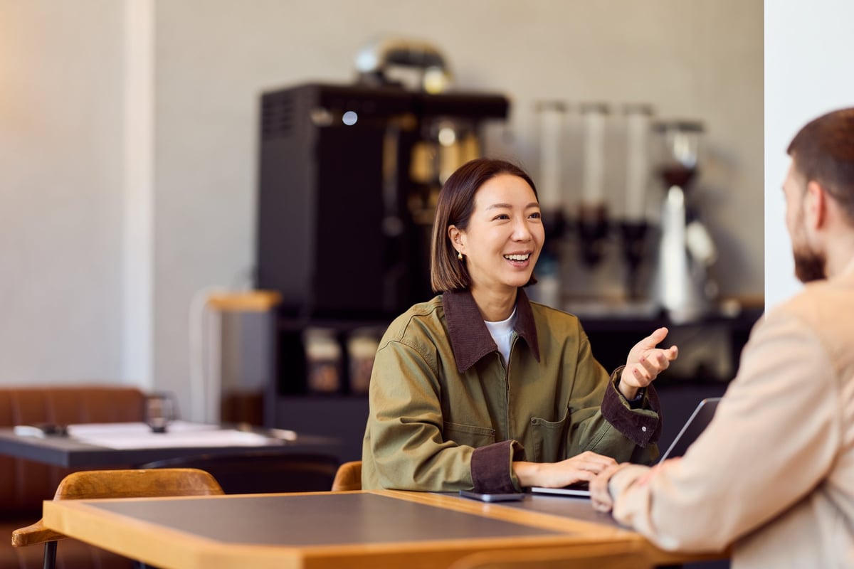 An Asian woman in a green jacket chats with a man across a table in a café, smiling and engaging in a relaxed business conversation